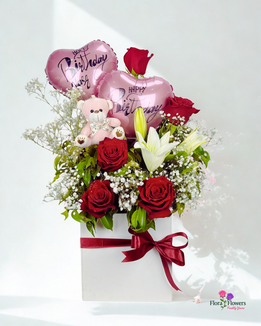Bouquet of red roses, white lilies, and balloons with a teddy bear on a white background, featuring the Flora & Faeries logo.