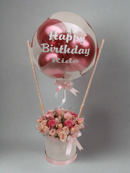 A pink and white floral arrangement with a 'Happy Birthday' balloon, roses, and a box of chocolates.