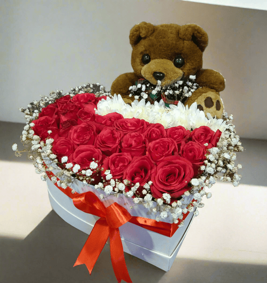 A basket of red roses with white flowers and a teddy bear, presented in a box with a ribbon.