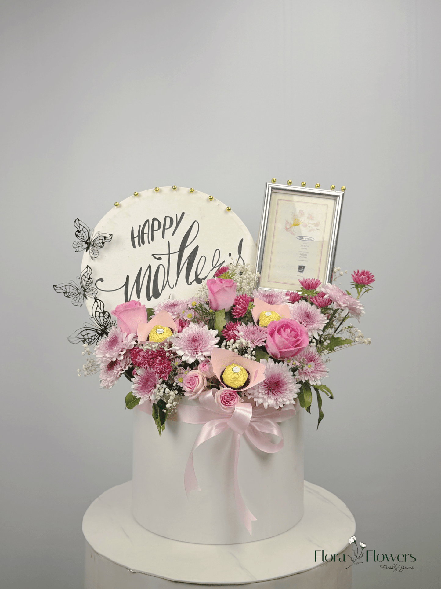 A round, white box with a floral arrangement on top, including pink roses, a 'Happy Mother's Day' sign, and a photo frame