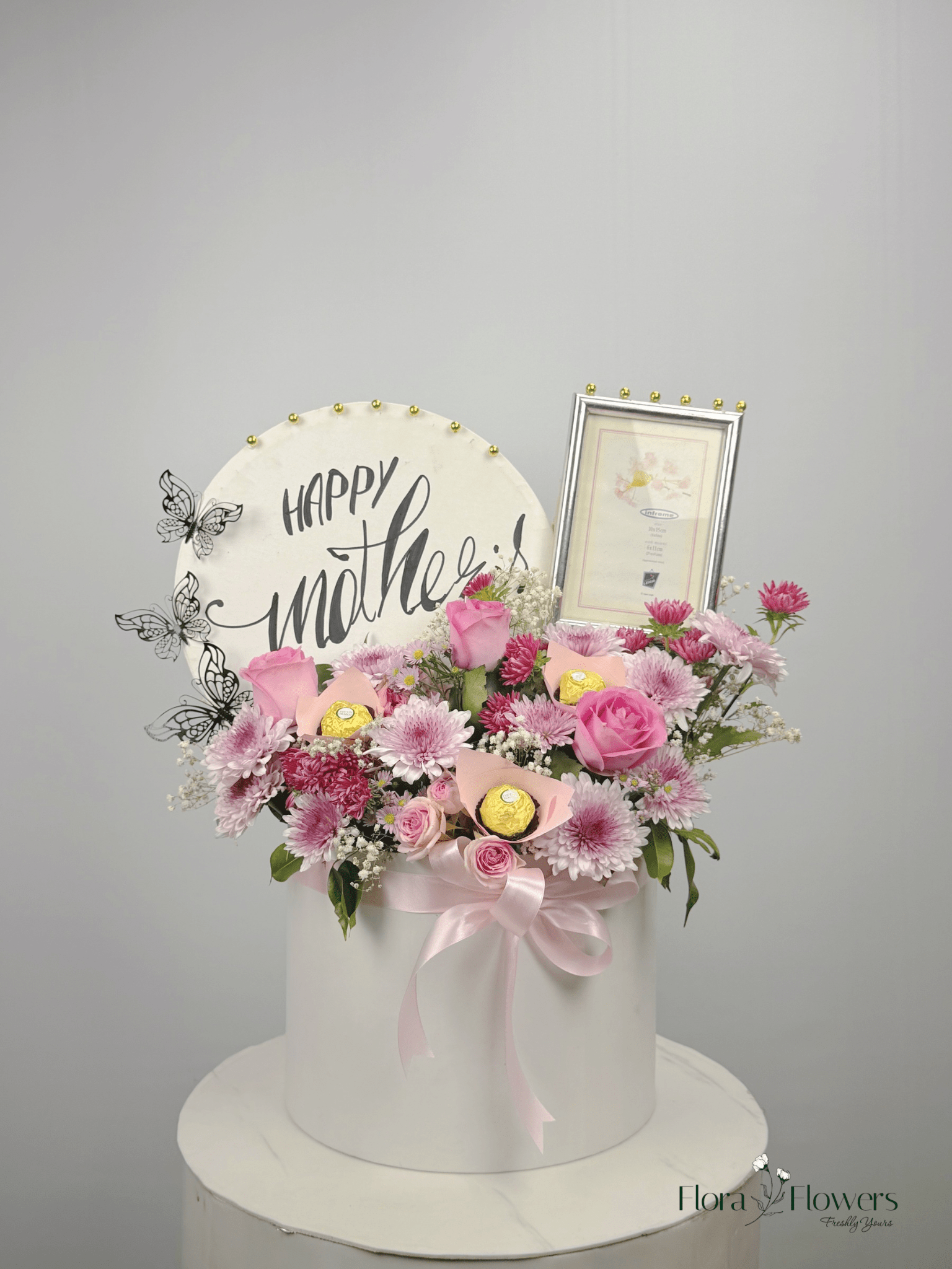 A round, white box with a floral arrangement on top, including pink roses, a 'Happy Mother's Day' sign, and a photo frame