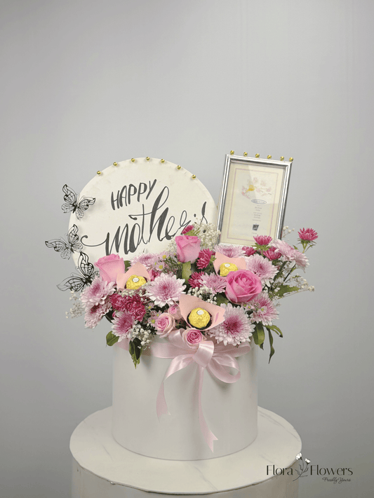 A round, white box with a floral arrangement on top, including pink roses, a 'Happy Mother's Day' sign, and a photo frame