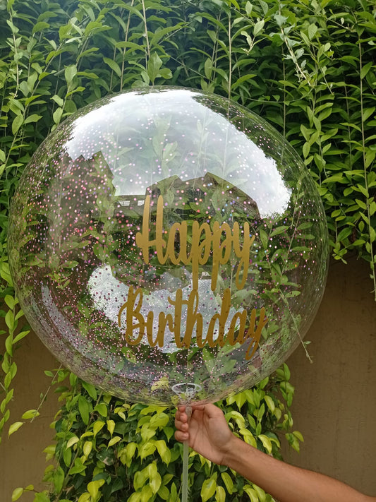 A hand holding a clear round balloon with 'Happy Birthday' text printed in gold.