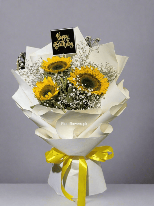A bouquet of three sunflowers and two Baby's Breath flowers, wrapped in white paper with a yellow ribbon, a 'Happy Birthday' tag, and additional decorative elements.