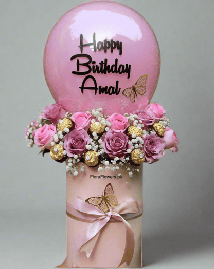 A birthday balloon with 'Happy Birthday Amal' printed on it, accompanied by an arrangement of pink roses, baby's breath, and chocolates.