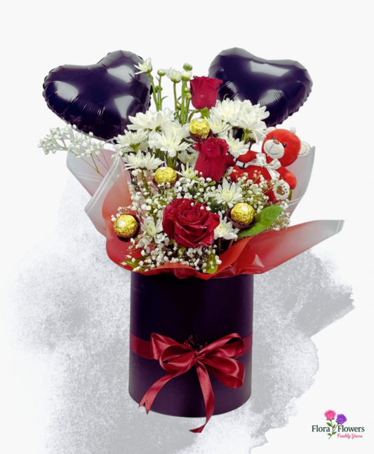 Floral arrangement with red roses, white flowers, and chocolate candies in a purple box with heart balloons on a white background.