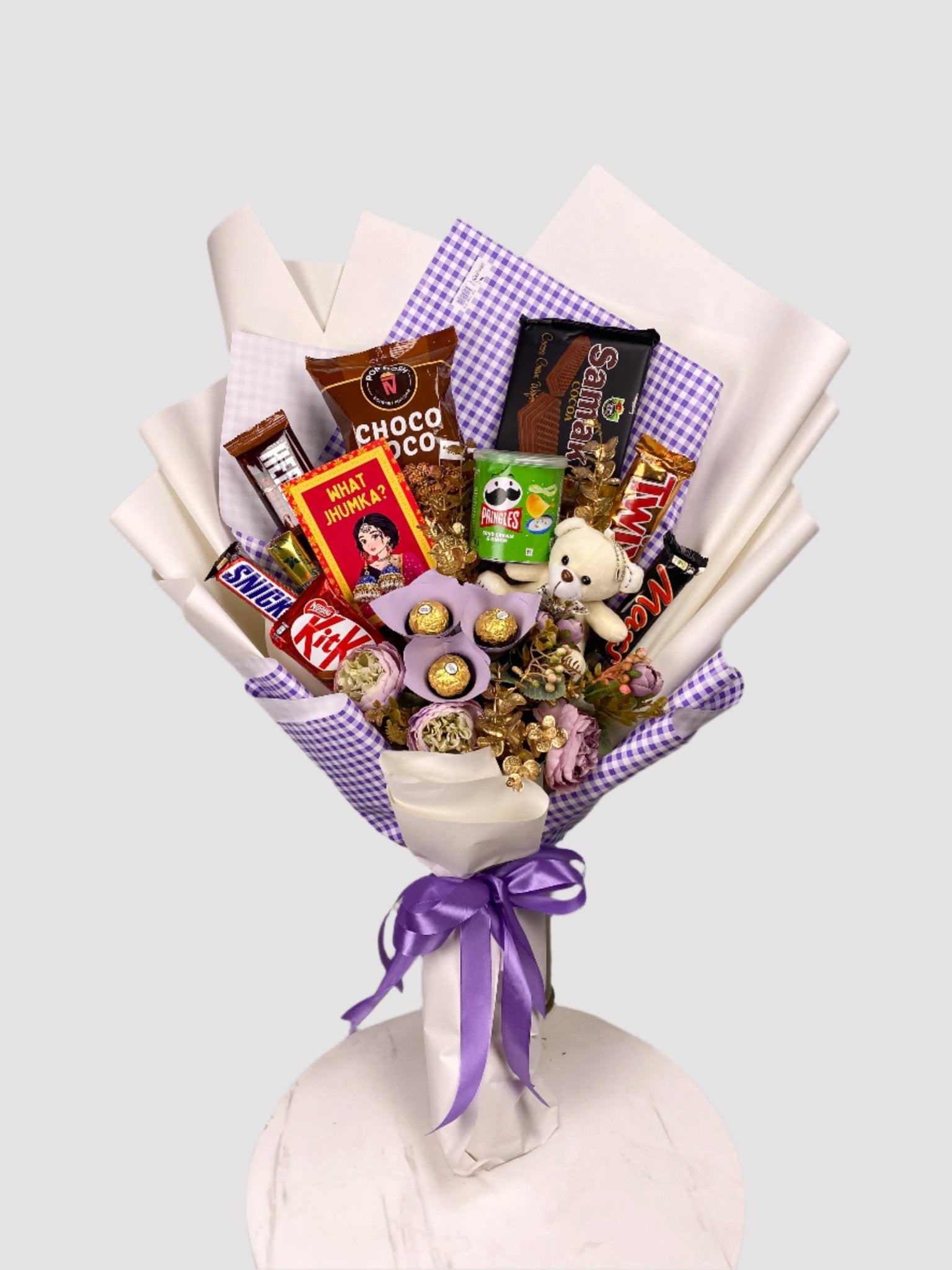 A bouquet arrangement made of various food items including chocolate bars and cookies, with decorative artificial flowers and a pair of earrings, all wrapped in checkered paper with a purple ribbon.
