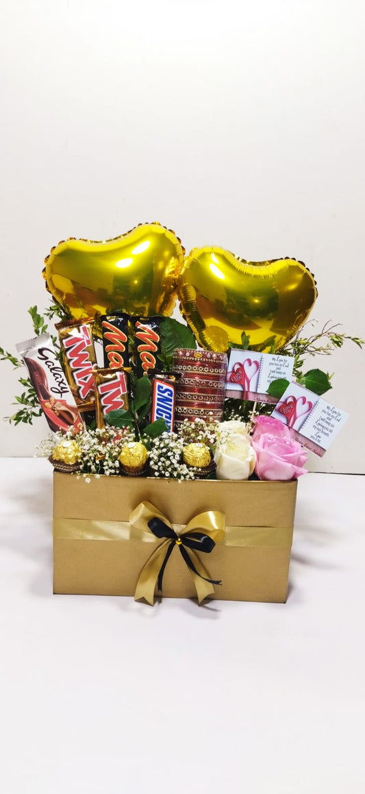 A gift basket with an assortment of chocolates, roses, and decorative balloons in golden yellow and pink colors, presented in a rectangular box with a bow on top.