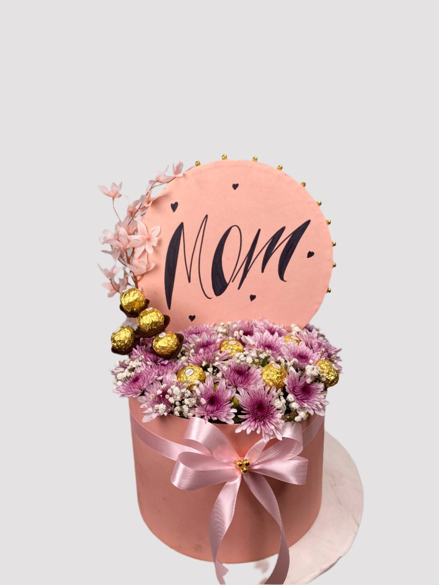 A round pink floral arrangement with the word 'Mom' on it, decorated with multicolored flowers and baby's breath, and tied with a ribbon.