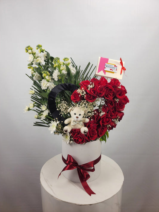 Heart-shaped flower arrangement with a teddy bear and a red ribbon on a white surface.