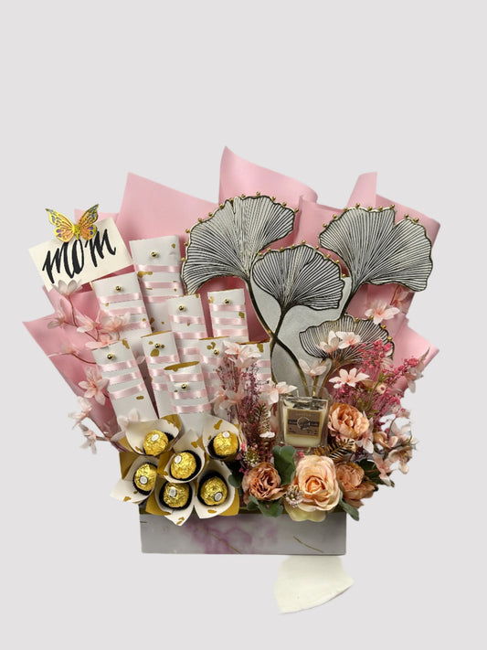 A gift arrangement featuring an assortment of chocolate candies including Ferrero Rochers, Twix, and KitKat, accompanied by artificial flowers and a candle, packaged in a box with pink tissue paper.