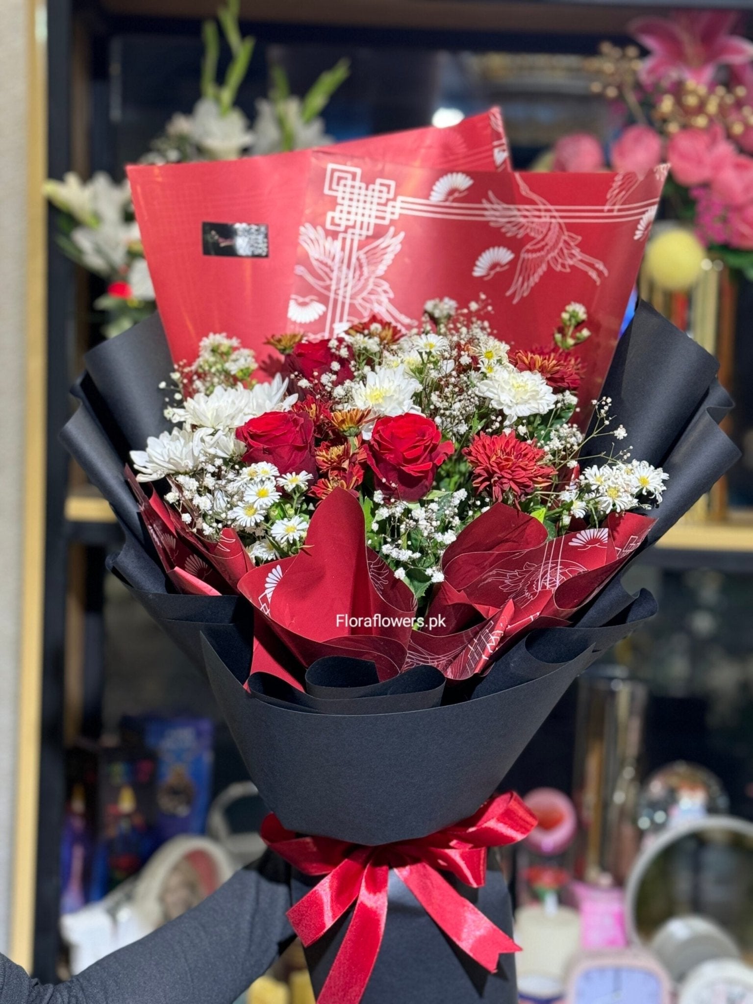 Bouquet of red and white flowers wrapped in black paper with a red ribbon, branded 'FloraFlowers.pk'.