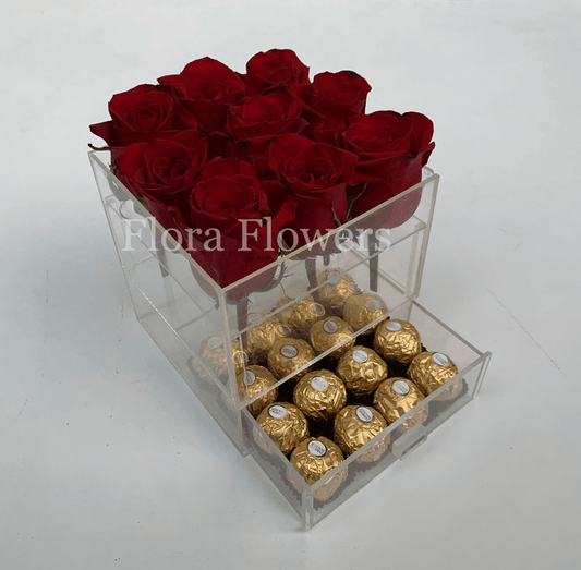 A clear acrylic box containing a dozen red roses arranged on top, with a layer of chocolate candies in a pattern below.