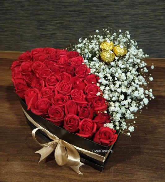 A heart-shaped basket arrangement featuring red roses and white flowers, with a gold bow and decorative elements.